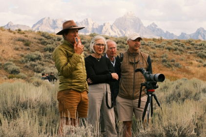 Four people observe mountains from a grassy field.